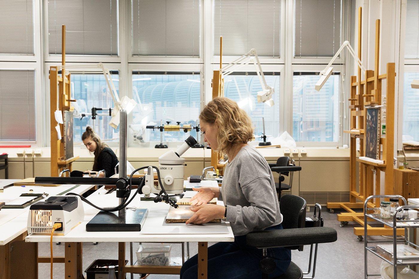 Zentral im Vordergrund sitzt eine Studentin an einem Arbeitstisch, das Foto zeigt sie von links im Profil. Vor ihr liegt ein kleines Gemälde, das sie gerade unter ein Mikroskop schiebt. Im Hintergrund links sieht man eine Studentin vor einem Gemälde auf einer Staffelei, dahinter die Fensterfront; rechts im Bild stehen weitere Staffeleien.