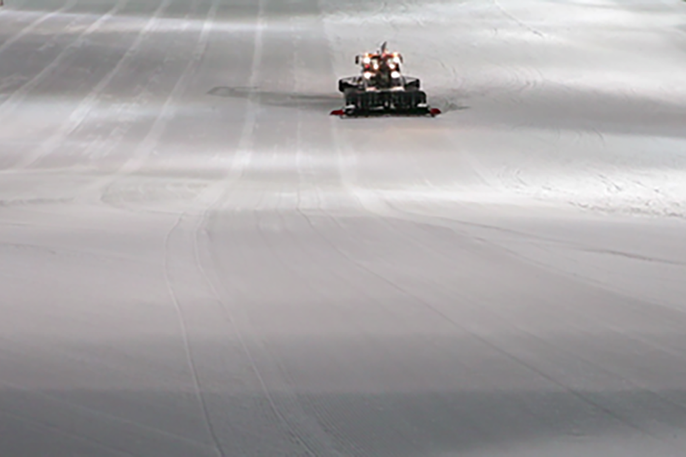 Videostill. Eine große weiße Fläche, eine geplante Schipiste, weiter hinten ist ein Fahrzeug zu erkennen, das die Piste präpariert.