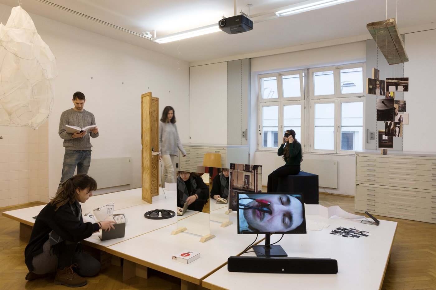 Mehrere Personen befinden sich in einem Raum, dessen Mitte ein niedriges Podest steht, auf dem mehrere Objekte aus unterschiedlichen Materialien stehen, im Vordergrund ein Monitor mit einem Gesicht, in dessen Nase Kabel stecken.