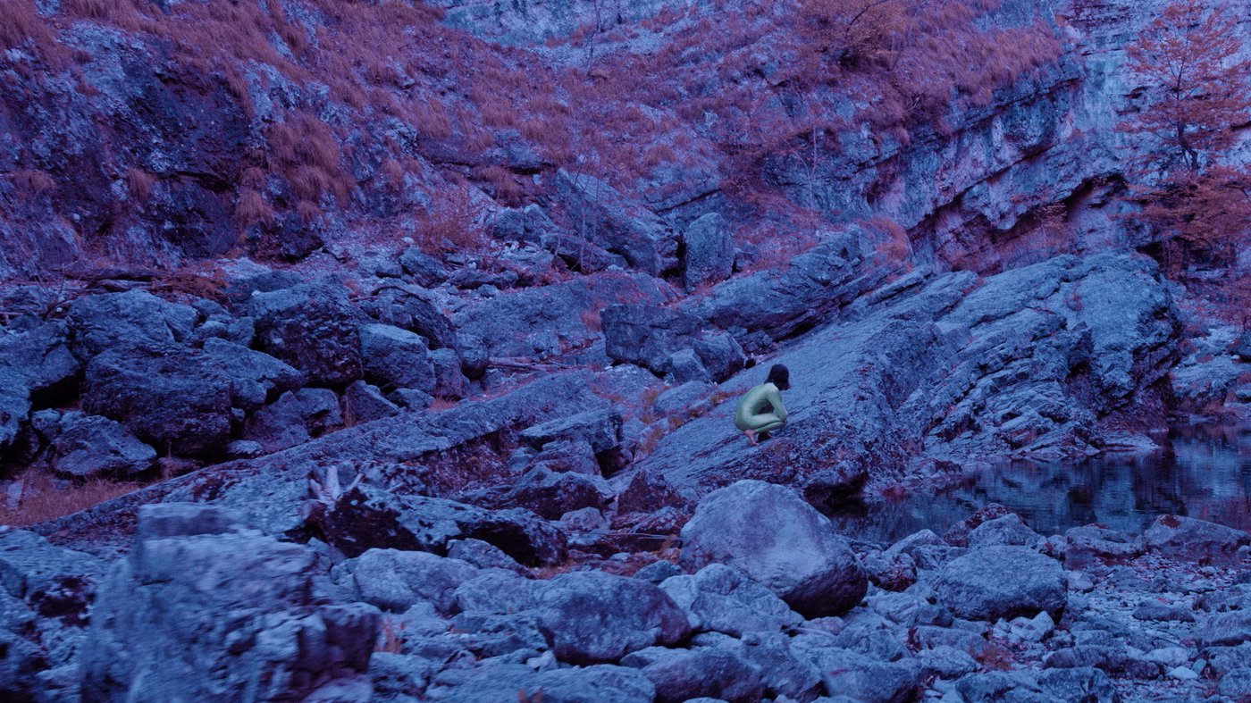 Eine violette Berglandschaft mit einer winzigen weiblichen kauernden Figur.