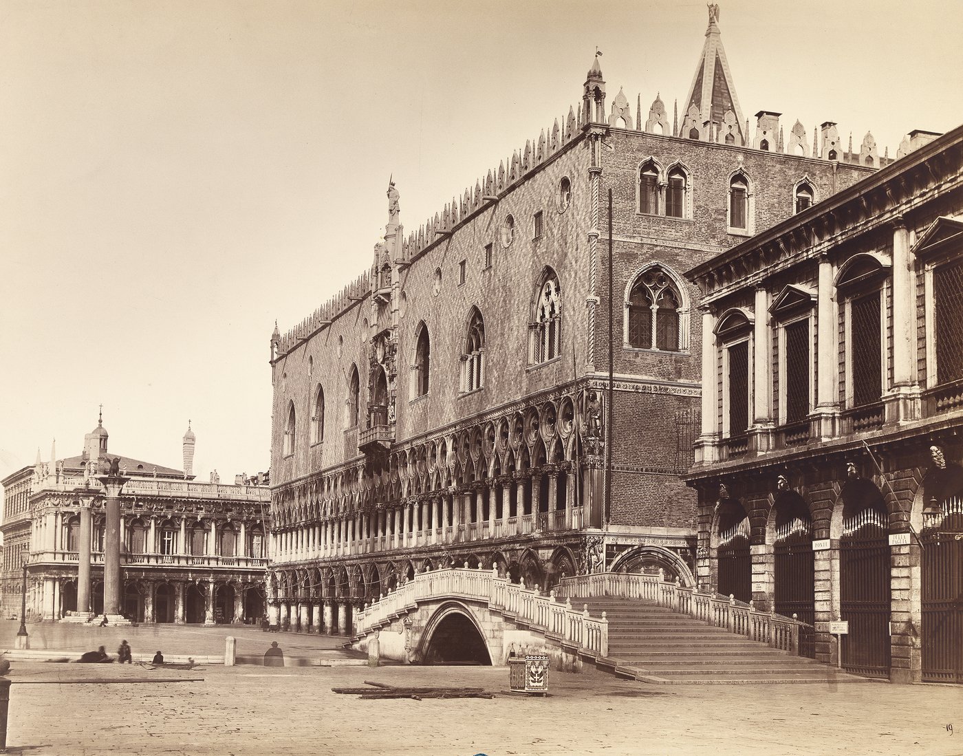 Eine Schwarzweißfotografie in Sepia, die den Dogenpalast und eine kleine Brücke abbildet. Die Architektur entspricht der Venezianischen Gotik und zeigt deshalb nach oben hin spitzzulaufende, unterteilte Fenster und Verzierungen, sowie einen Loggia anmutenden Durchgang, der von Säulen und Pfeilern getragen wird. Auf der linken Bildseite sind vereinzelte Personen leicht verschwommen zu erkennen.