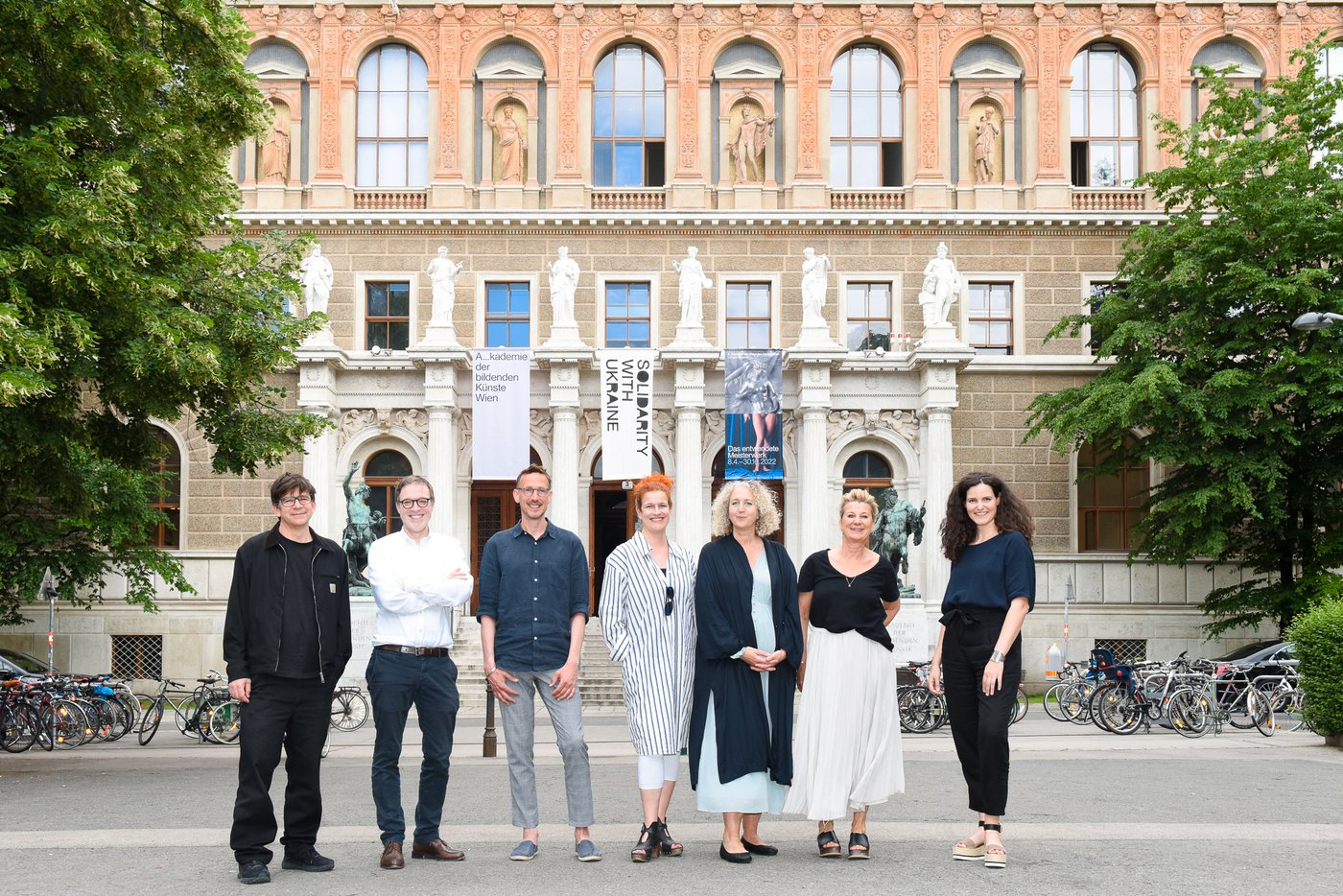 Foto von 7 Personen aufgereiht vor dem Akademiegebäude lächeln in die Kamera, im Hintergrund Bäume