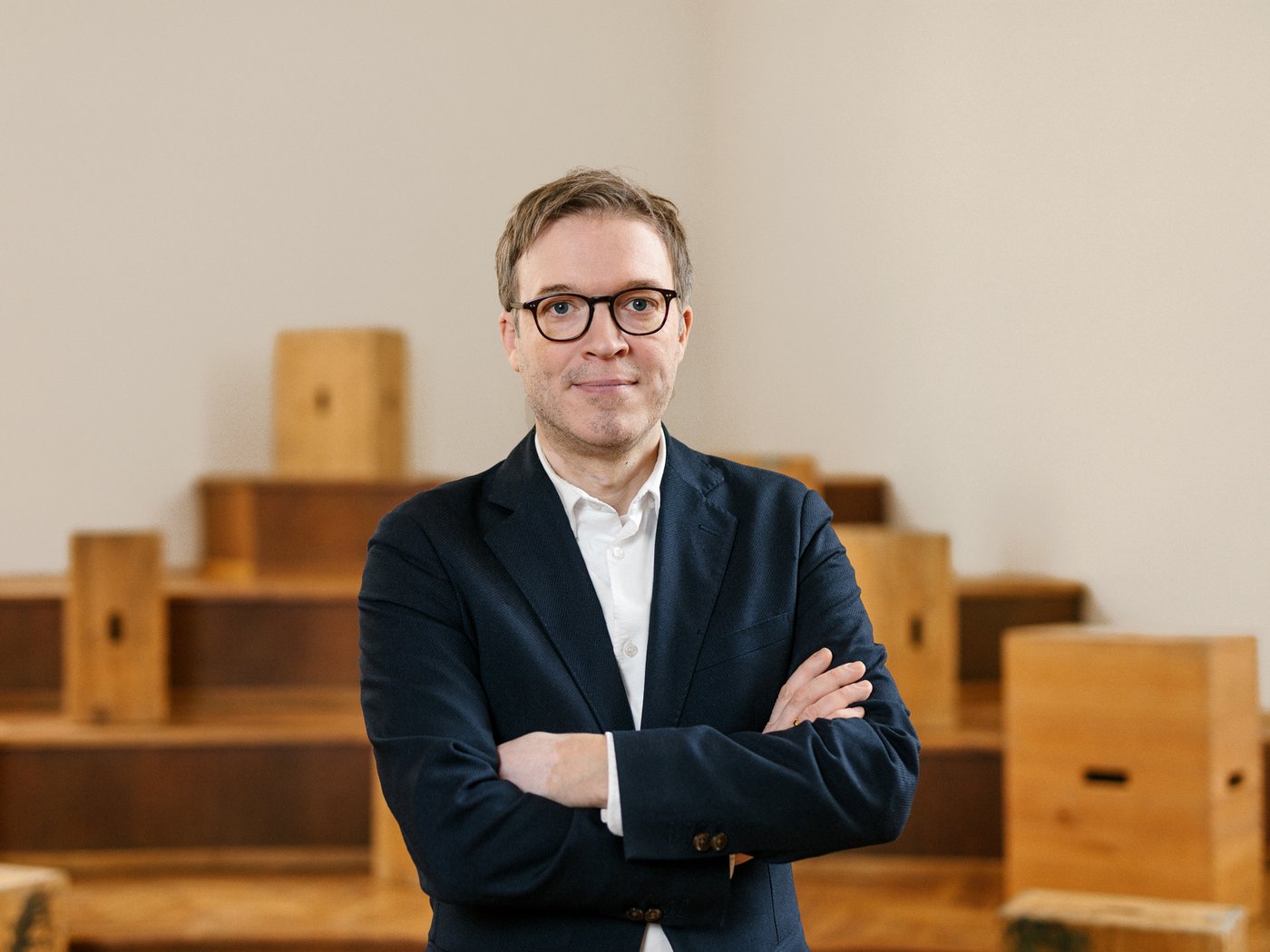 Mann in einem dunkelblauen Blazer mit einem weißen Hemd steht mit verschränkten Armen vor einer weißen Wand, am Boden unscharf hölzerne Objekte und Stufen