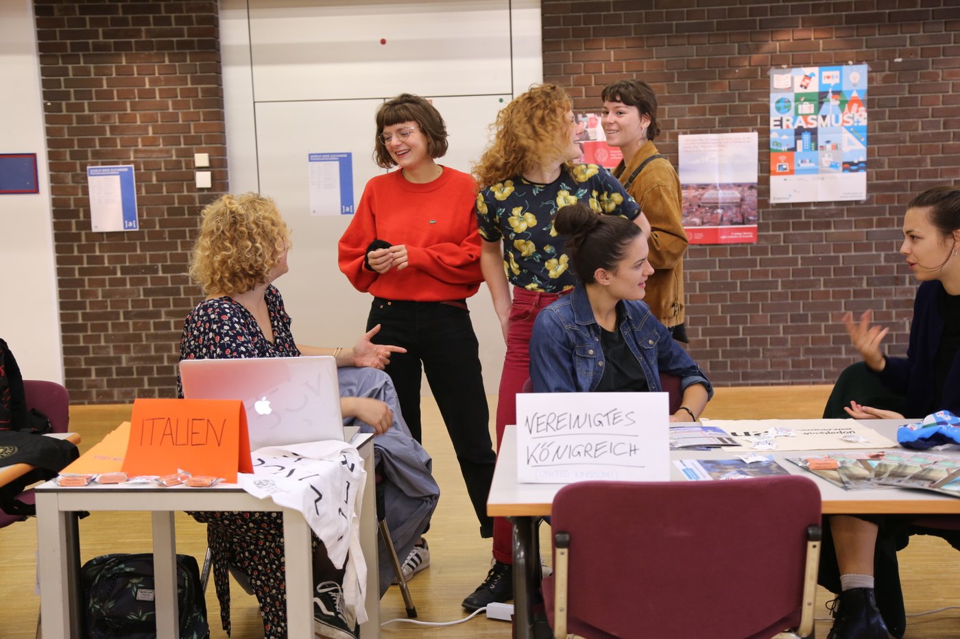 Man sieht Incoming Austauschstudierendeaus Italien und dem vereinigten Königreich im lebhaften Gespräch mit Studierenden, die sich für einen Auslandsaufenthalt interessieren. An der Rückwand hängt ein Plakat, welches auf das Mobilitätsprogramm Erasmus plus aufmerksam macht.