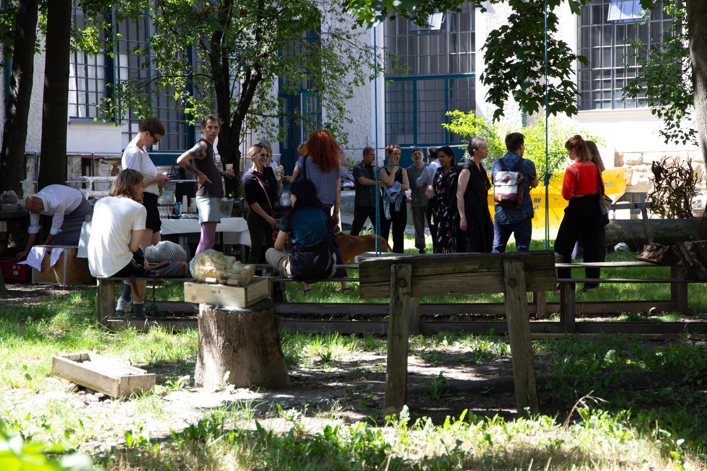 Menschen stehen bei einer feierlichen Zusammenkunft in einen grünen Innenhof. Es ist Sommer, Bänke und ein Tisch mit Getränken wurden aufgestellt. Dazwischen liegen Gerätschaften und Kisten, Teile von Skulpturen. Im Hintergrund sieht man die großen Atelierfenster des Gebäudes.