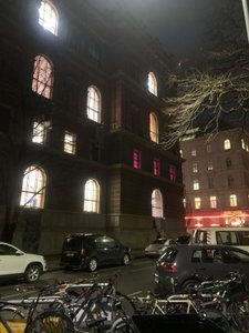 Das Bild zeigt das Hauptgebäude der Akademie der bildenden Künste Wien am Schillerplatz bei Nacht. Es ist ein historisches Gebäude mit mehreren großen, erleuchteten Fenstern. Besonders auffällig sind drei Fenster im mittleren Bereich, die in einem kräftigen Pink leuchten. Vor dem Gebäude befinden sich geparkte Autos und zahlreiche Fahrräder. Im Hintergrund sieht man ein weiteres beleuchtetes Gebäude.