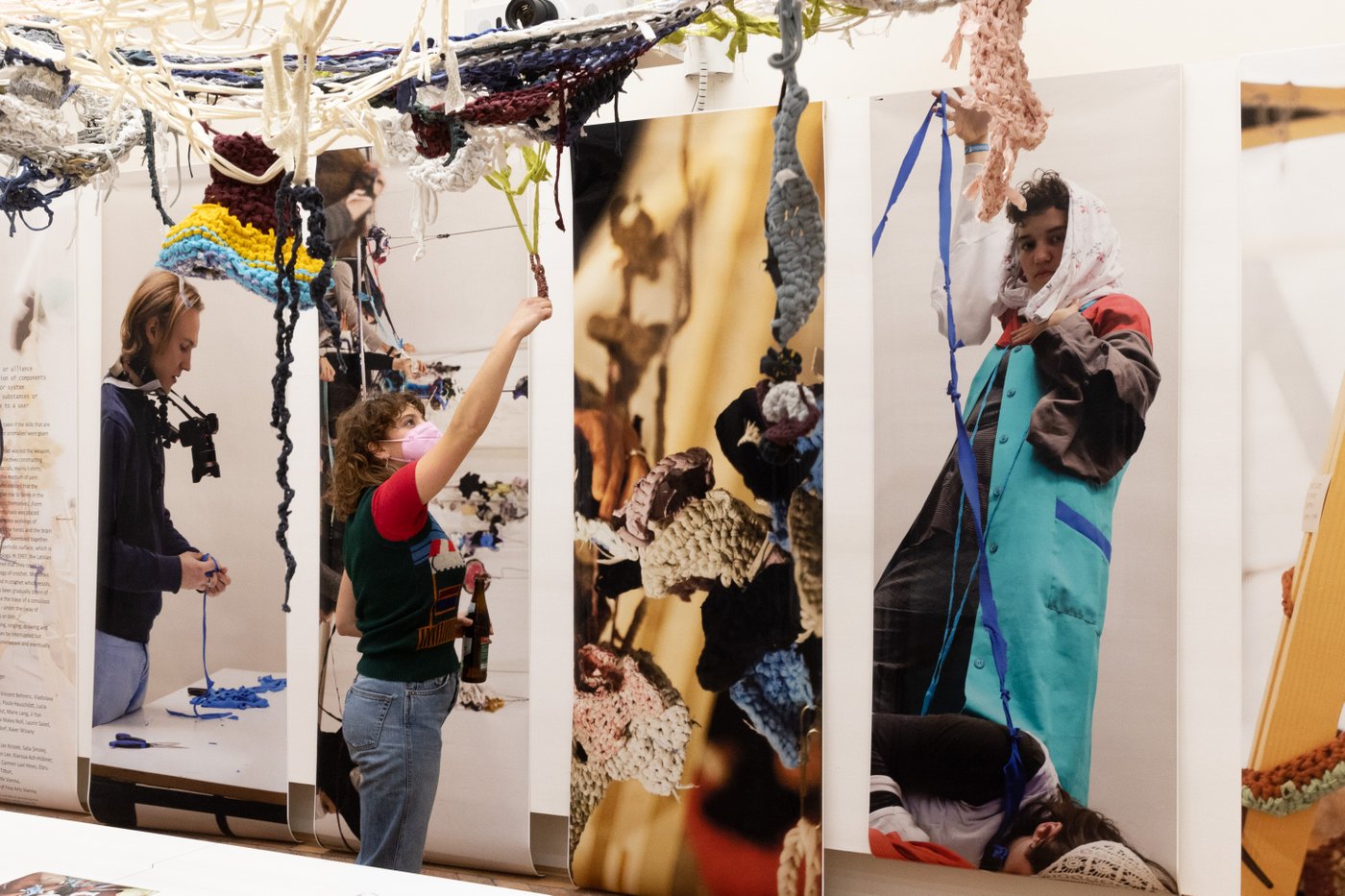 Colourful threads of fabric hang from the ceiling, colourful posters with people on them hang on the wall, a person stands in the room and stretches towards the threads of fabric on the ceiling.