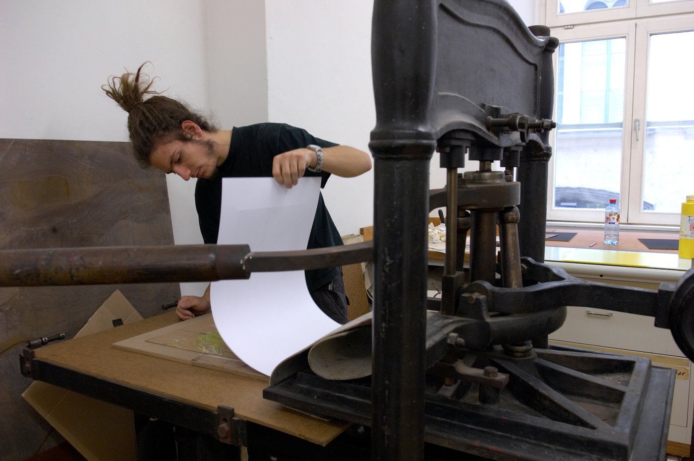 A student stands at a large heavy printing machine and is checking the result by lifting up the folded sheet.