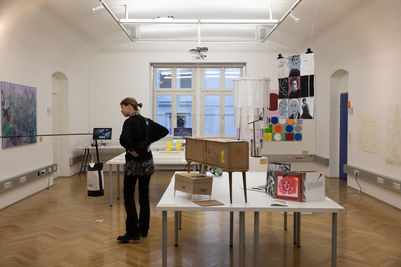 Presentation situation with two large tables on which you can see various objects. From the ceiling hangs a textile work with portraits of women, next to it a kind of lamp, a visitor looks at a picture on the wall.