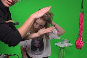The image shows a still from the video Green Scream with G-udit, in the foreground we see a clapping hand, a woman in the background runs her hand through her hair.