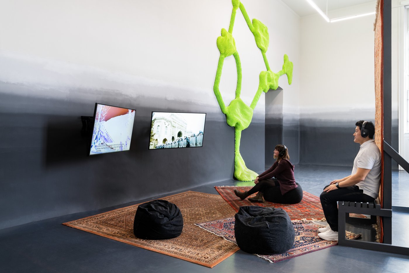 Two people are sitting on carpets in front of 2 screens. In the background you can see a green structure.