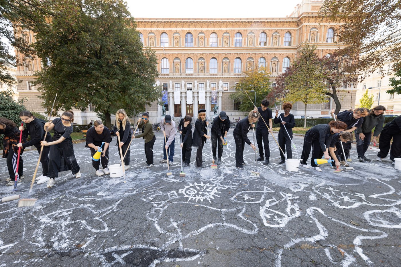 Several students working together to draw a chalk drawing with white paint and brooms on Schillerplatz.