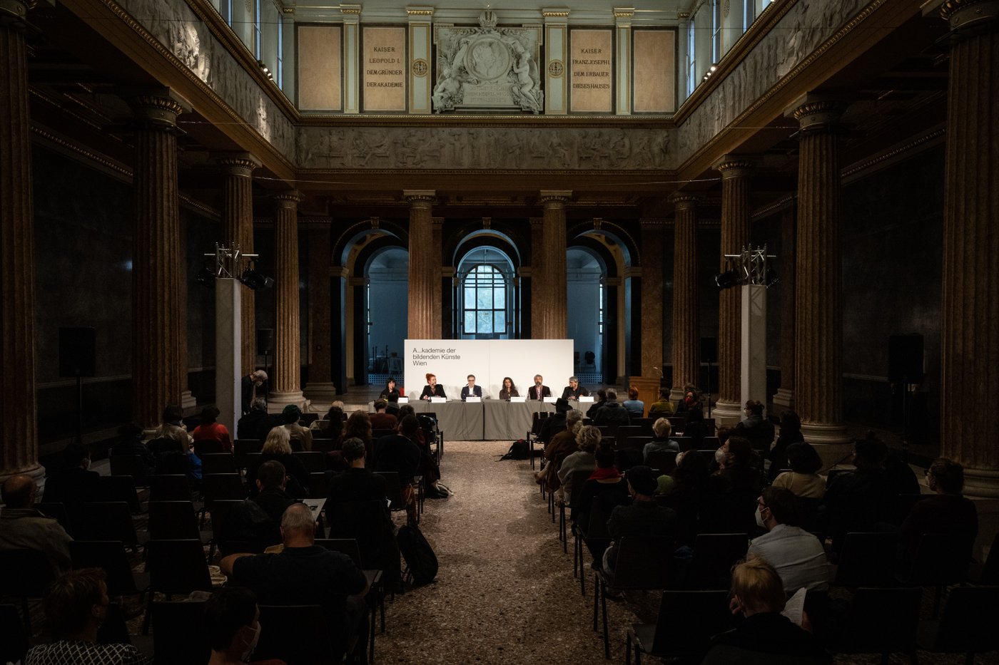 Five people are seated at a long table in a historical room enclosed by columns, with an audience in front of them in semi-darkness.