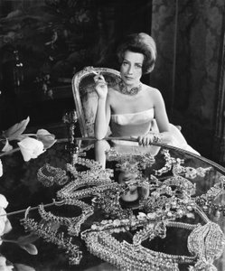 Black and white photograph of a woman in a white dress sitting at a round table on which jewellery is laid out.