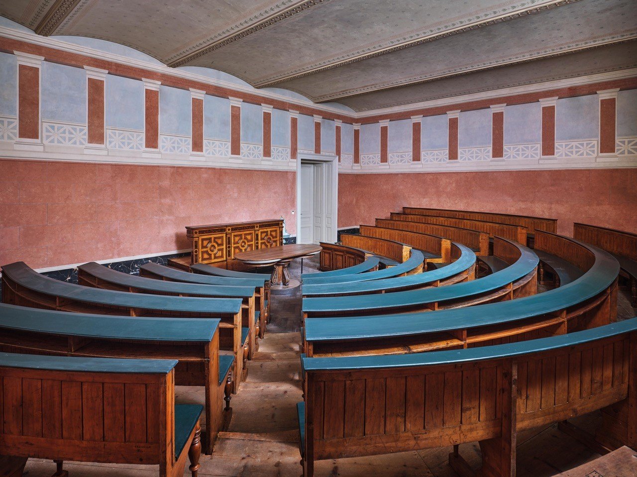 Photograph of a lecture hall in which the rows of wooden benches have been arranged in a stepped semicircle, with a table and lectern in the center, the walls painted in terracotta colors with illusionistic columns interrupted by a white railing.