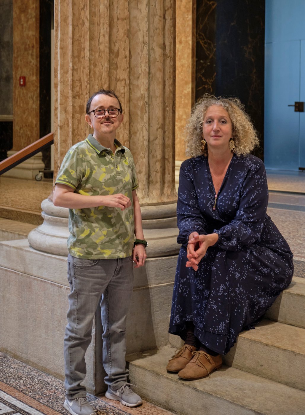 Photo of two people framed by Greek columns, one person standing, the other sitting on a step