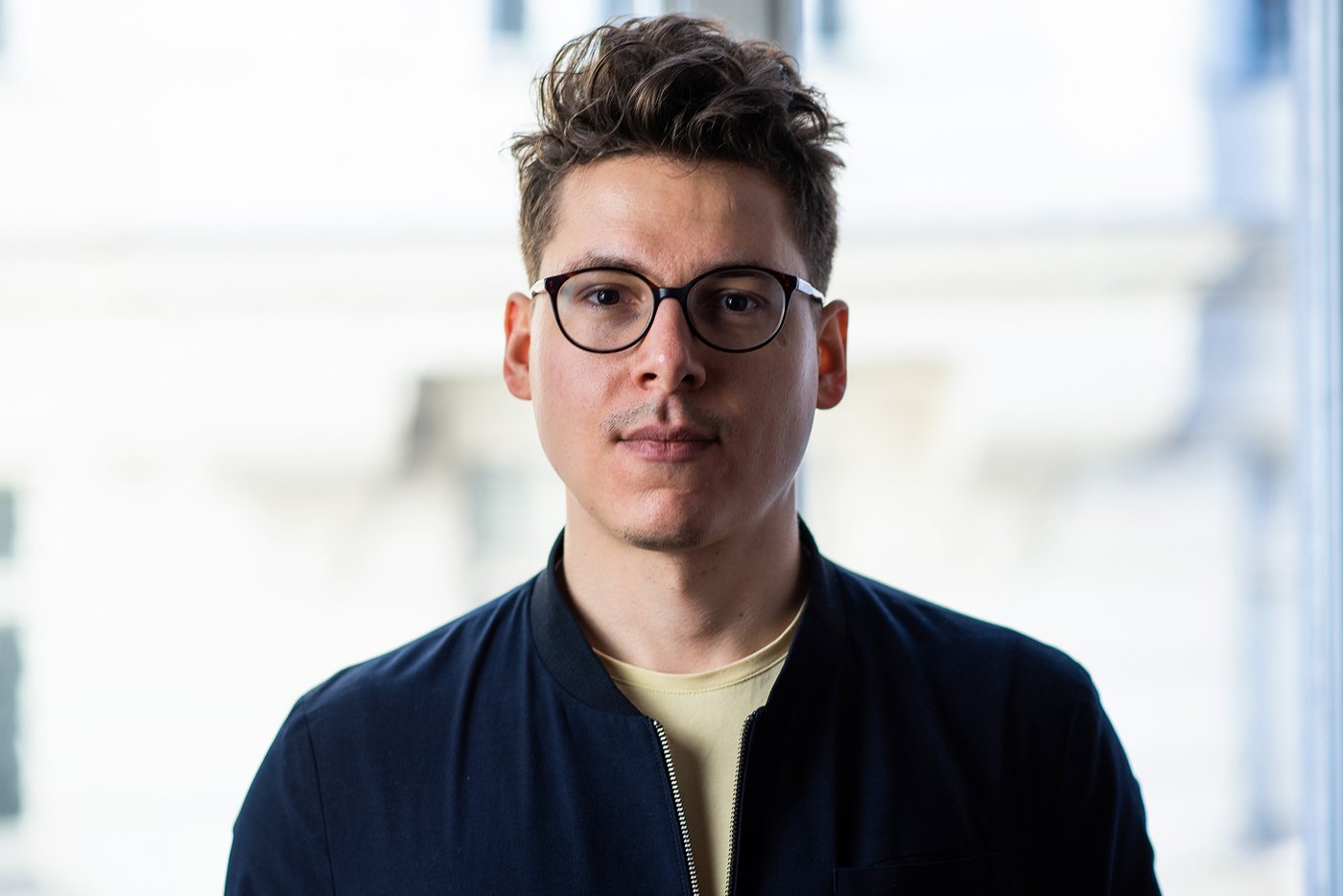 The color photograph in landscape format shows a chest portrait of Lukas Meschik. The young author wears large glasses, his brown hair is cut short at the sides and his longer top hair is combed upwards. He is wearing a light-colored T-shirt with a dark blouson over it.