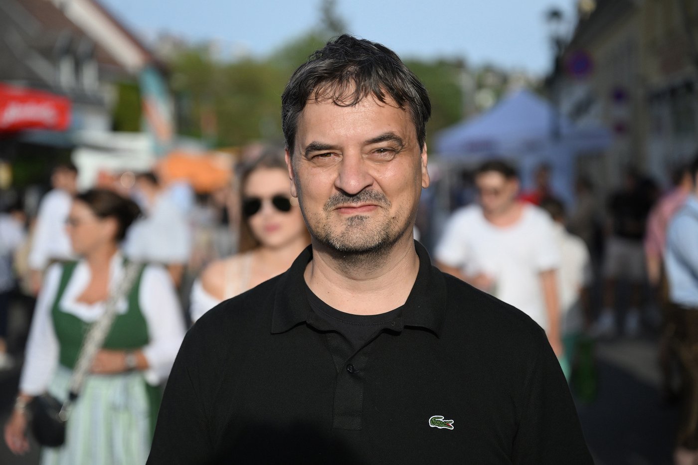 The color photograph in landscape format shows a chest portrait of Peter Waldeck. The friendly-looking man in his prime with a full head of dark hair has his left eyebrow slightly raised and is wearing a black long-sleeved T-shirt. A busy street can be seen blurred in the background.